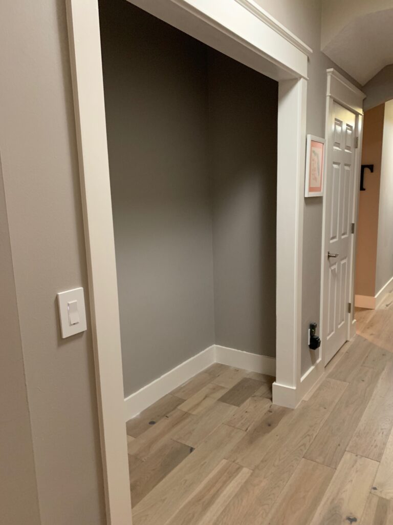 Right side of a closet that missing doors and the shelving, with white trim and wood floors, in a hallway.