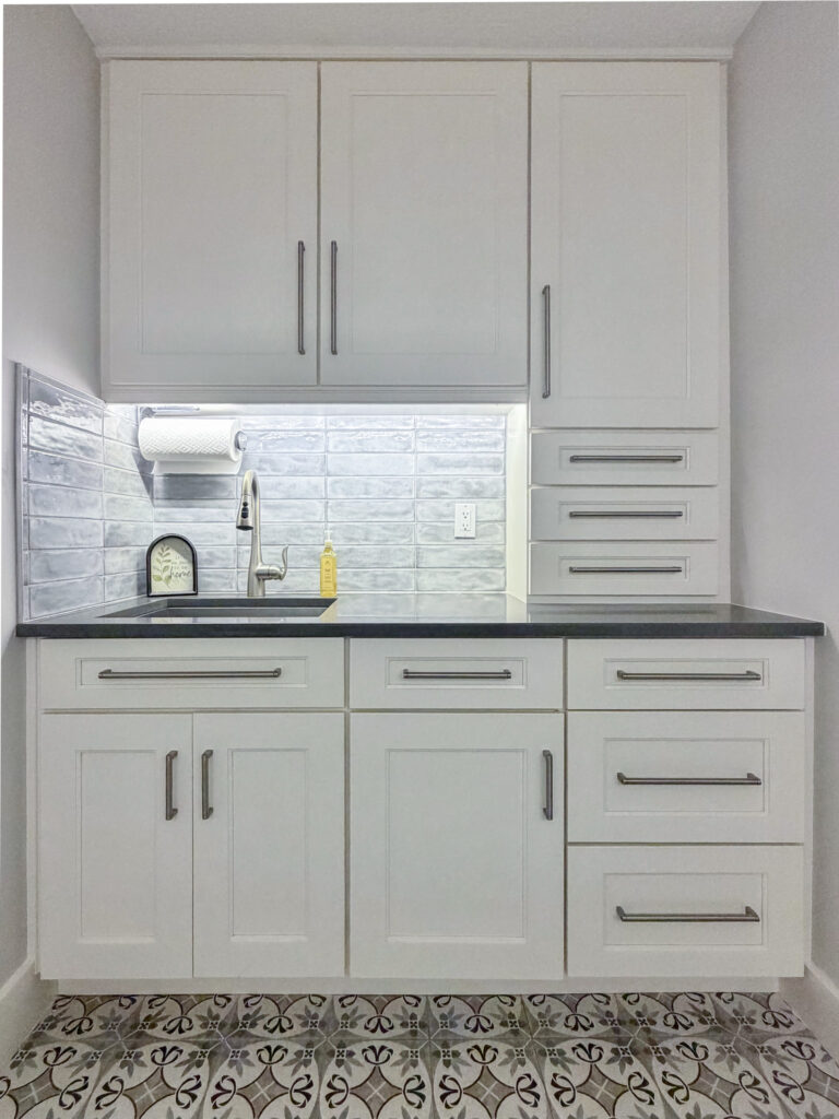 Laundry room after a renovation with washer and dryer, custom white cabinets, and utility sink with patterned tile floor, subway tile backsplash, and quartz countertops.