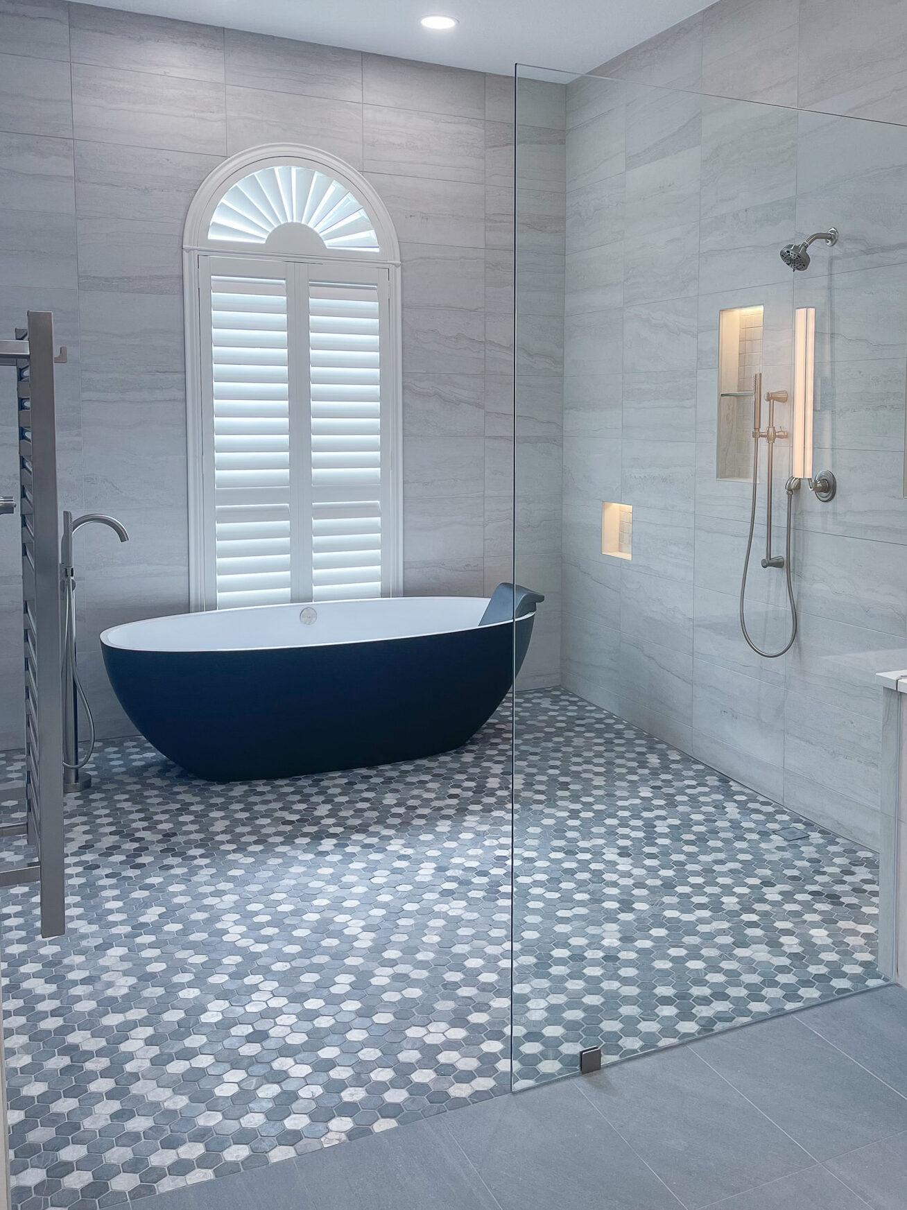 Zero-entry wet room with freestanding tub, illuminated shower niches, hex tile flooring, towel warmer, and custom wood vanity.